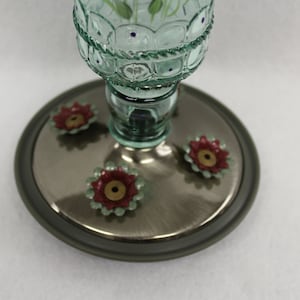 Puede incluir: Un comedero para colibr&iacute;es de vidrio verde con una base plateada y cuatro puertos de alimentaci&oacute;n en forma de flores rojas y verdes.