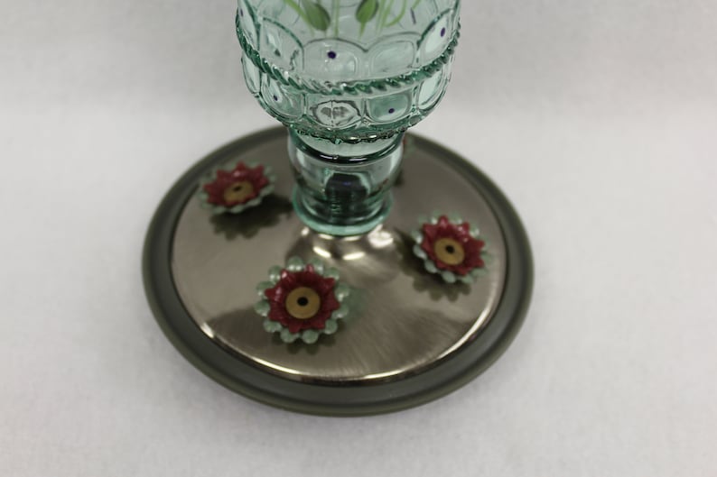 Puede incluir: Un comedero para colibr&iacute;es de vidrio verde con una base plateada y cuatro puertos de alimentaci&oacute;n en forma de flores rojas y verdes.