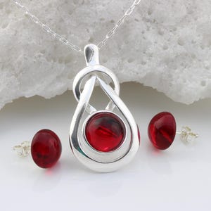 May include: A silver pendant necklace and matching stud earrings set. The pendant has a teardrop design with a red gemstone centre. The earrings also have red gemstones. The jewellery set is displayed on a white surface.