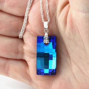 May include: A rectangular blue crystal pendant necklace with a silver chain. The pendant is a vibrant blue with a faceted design, reflecting light. The necklace is a delicate piece of jewellery.
