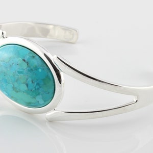 Pode incluir: Um bracelete de prata com uma pedra oval turquesa engastada no centro. O bracelete tem um design simples e moderno.