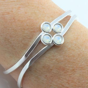 May include: A silver cuff bracelet featuring four opalescent stones in a square arrangement. The bracelet has a minimalist design with two parallel bands that curve around the wrist. The stones have a pearly, iridescent quality, reflecting light. A stylish piece of jewellery.