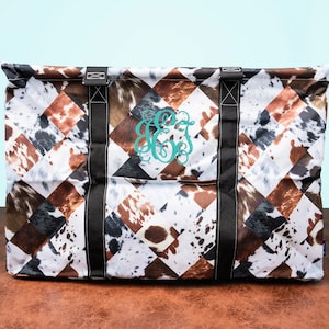 May include: Large tote bag with a cowhide pattern in brown, white, and black diamond shapes. The bag has black straps and a teal monogram. The bag is designed for carrying items.