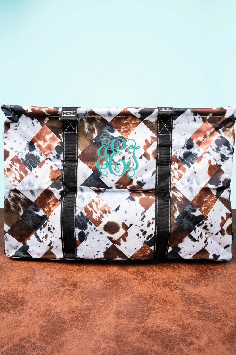 May include: Large tote bag with a cowhide pattern in brown, white, and black diamond shapes. The bag has black straps and a teal monogram. The bag is designed for carrying items.