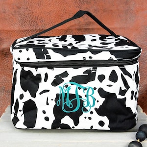 May include: A black and white cow print lunch bag with a black zipper and handle. The bag has a teal monogram of the letters MJB. The bag is sitting on a gray wooden crate.
