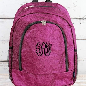 May include: A magenta glitter backpack with black trim and a black zipper. The backpack has a large front pocket with a black zipper and a black monogram. Mesh pockets are on the sides.
