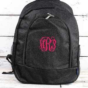 May include: Black backpack with a pink monogram. The backpack has a zippered main compartment, a front pocket, and side mesh pockets. The monogram is in a decorative script font. The backpack is made of a textured material.