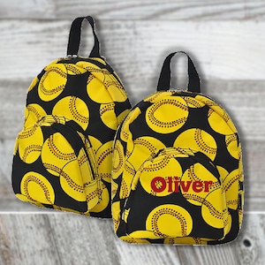 May include: Two black and yellow softball-themed backpacks. The backpacks feature a pattern of yellow softballs on a black background. One backpack has the name "Oliver" embroidered in red on the front pocket. Both have black handles and zippers.