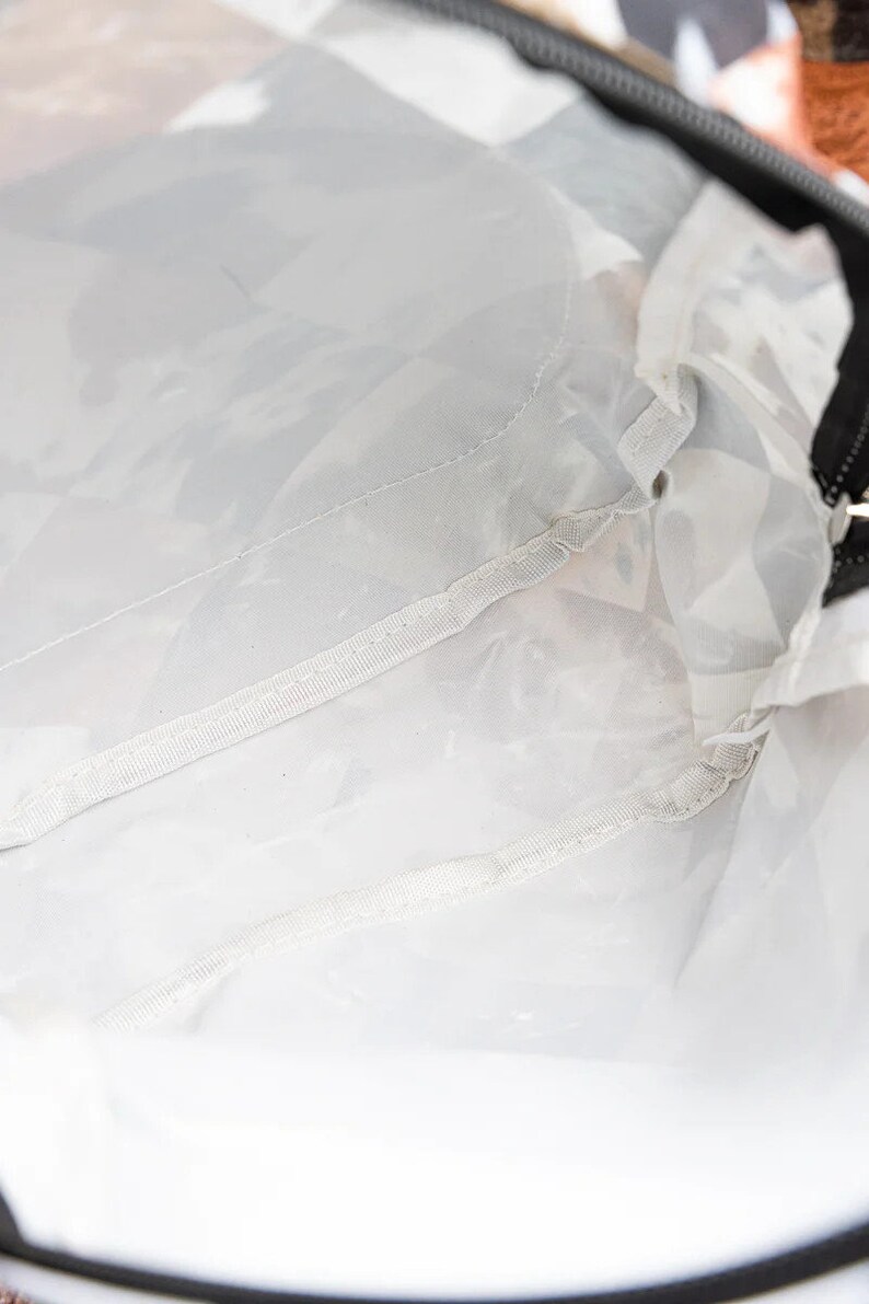 May include: A clear, white, and gray bag interior with a black zipper. The bag's interior is made of a translucent material, with seams and stitching visible. The bag's design is simple and functional.