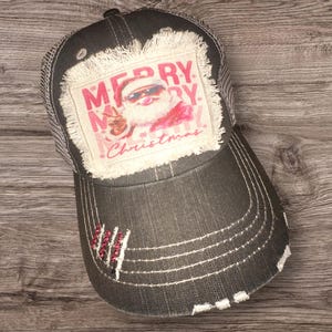 May include: Distressed black baseball cap with a mesh back and a frayed patch. The patch features a pink and white graphic with the words "MERRY CHRISTMAS". The brim has white stitching and a decorative red embellishment.