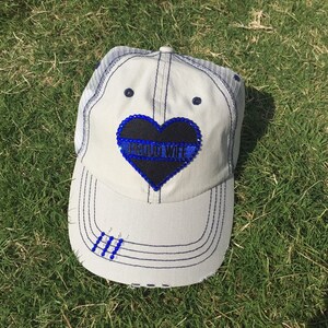 May include: White trucker hat with a black heart-shaped patch that says "PROUD WIFE" in blue with a thin blue line across the heart. The hat has a blue mesh back and a blue stitched brim.