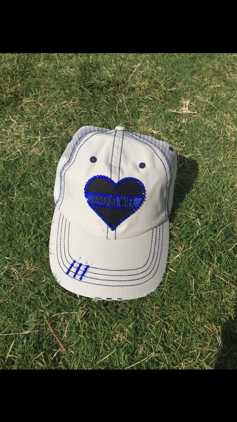 May include: White trucker hat with a black heart-shaped patch that says "PROUD WIFE" in blue with a thin blue line across the heart. The hat has a blue mesh back and a blue stitched brim.