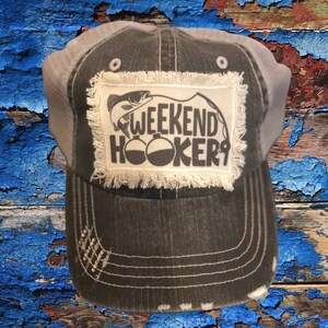May include: A black and white distressed trucker hat with a white patch that says "Weekend Hooker" and a graphic of a fish with a fishing bobber.