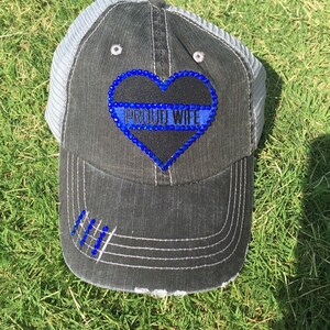May include: A black and gray baseball cap with a blue and white heart-shaped patch that says "PROUD WIFE" in rhinestones. The cap has a mesh back and a distressed look.