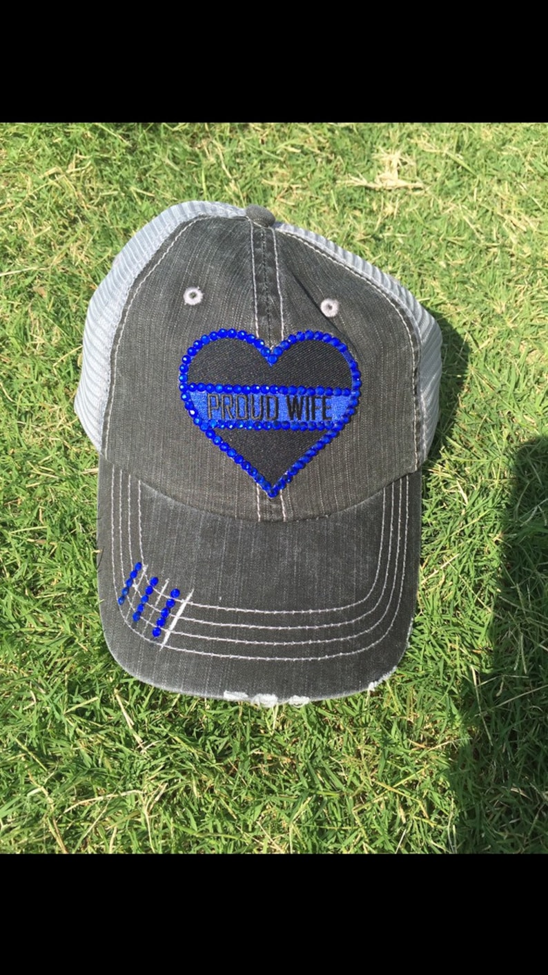 May include: A black and gray baseball cap with a blue and white heart-shaped patch that says "PROUD WIFE" in rhinestones. The cap has a mesh back and a distressed look.