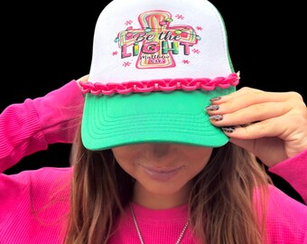 May include: A white and green trucker hat with the text "Be the Light" and a cross design. The hat has a pink chain detail on the brim. The hat is being held up by a person wearing a pink long-sleeve shirt.