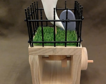 May include: A wooden Halloween-themed toy featuring a white ghost figure rising from a miniature graveyard. The graveyard includes a black fence, a gray tombstone, and green turf. The toy has a wooden crank and a light brown base.