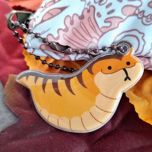 May include: A small, orange and brown, cartoon-style, snake-like charm with a smiling face. The charm is attached to a brown chain and is hanging from a white and pink patterned bag.