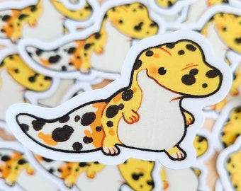 May include: A close-up of a pile of stickers featuring cartoon leopard geckos. The geckos are yellow, orange, and black with white bellies. The stickers are on a wooden surface.