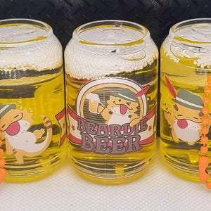 May include: Three miniature beer cans with orange chains. Each can has a cartoon bearded dragon wearing a hat and holding a beer mug. The middle can says "Beardie Beer" on it.