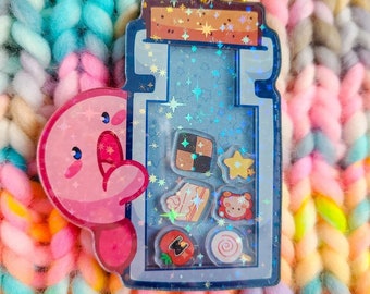 May include: A blue and pink glitter charm with a pink character peeking out of a bottle filled with colorful charms. The charm is attached to a gold star-shaped keychain.
