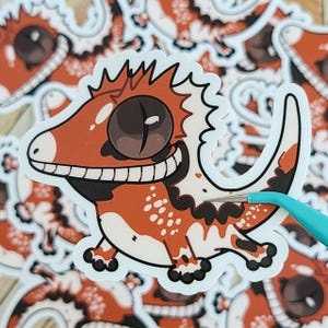 Puede incluir: Una pegatina con una ilustración de dibujos animados de un gecko rojo y blanco con una sonrisa de dientes y un gran ojo marrón. El gecko está de pie sobre sus patas traseras y tiene una cola larga y enroscada.