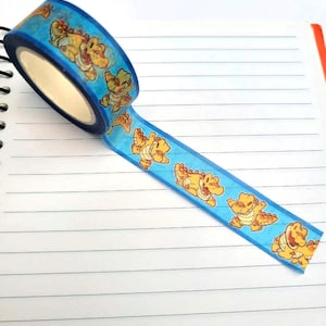 May include: A roll of blue washi tape with a repeating pattern of yellow cartoon dinosaurs. The tape is partially unrolled and is on a lined notebook.