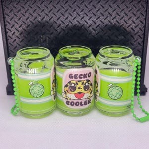 May include: Three green and white cans with a gecko design and the text "Gecko Cooler". Each can has a green chain attached to the top.