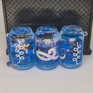 Op de afbeelding: Drie blauwe hars sleutelhangers in de vorm van een blikje. Elke sleutelhanger heeft een ontwerp met de woorden "Blue Raspberry Rattler" en decoratieve elementen. De sleutelhangers hebben een blauwe kralenketting en een zilverkleurige ring.
