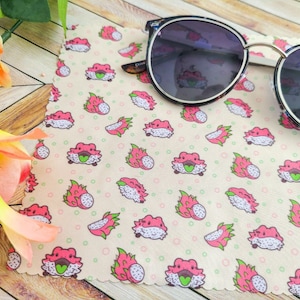 May include: A microfiber cleaning cloth with a white background and a repeating pattern of pink dragon fruit with green leaves. The cloth has a scalloped edge. A pair of round sunglasses with black frames and dark lenses is lying on top of the cloth.