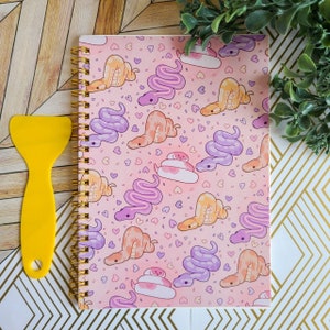 May include: A pink spiral bound notebook with a colorful pattern of snakes and hearts. The notebook has gold rings and a white background.