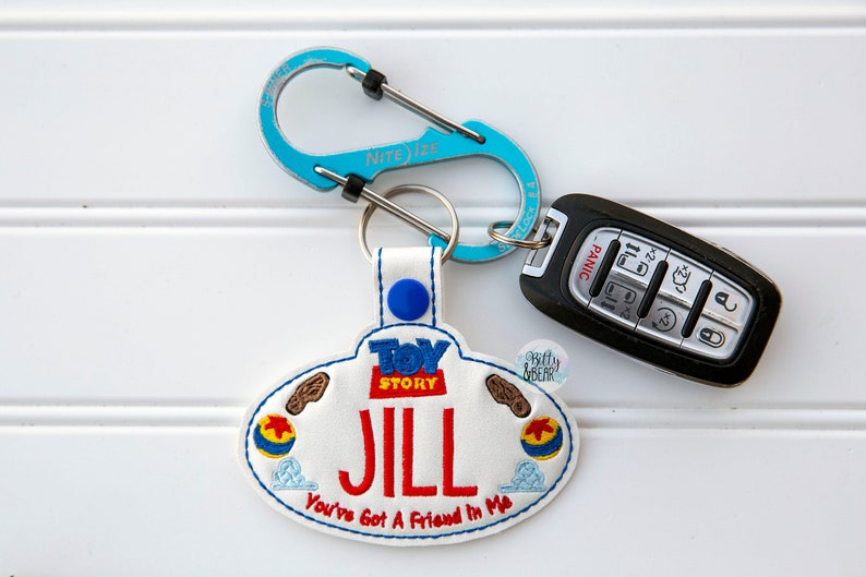Może przedstawiać: Biały brelok do kluczy z niebieskim i czerwonym logo Toy Story, imieniem "JILL" wyszytym na czerwono i frazą "You've Got A Friend In Me" wyszytą na niebiesko. Brelok ma niebieski karabińczyk i czarny kluczyk do samochodu.