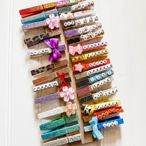 May include: A collection of wooden clothespins decorated with glitter, rhinestones, and small charms. Each clothespin has a different word or phrase spelled out with colorful letter beads, including "Fly High", "Shine", "Fierce", and "Awesome".