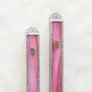 Puede incluir: Dos estuches de mezuzá de vidrio rosa con marcos plateados y letras hebreas. Los estuches están colgados en una pared blanca.
