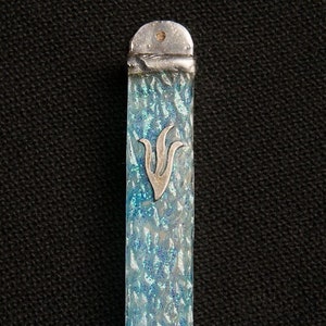Peut inclure: Un étui de mezouzah rectangulaire vertical avec un motif texturé bleu et transparent, parsemé de paillettes. L'étui a des capuchons en métal argenté à chaque extrémité et un emblème argenté au centre.