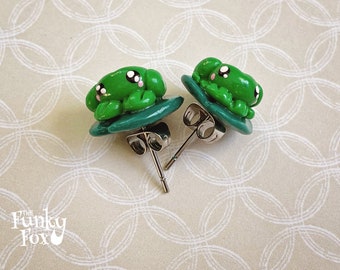 May include: A pair of green frog-shaped stud earrings with white eyes and a silver post. The earrings are made of polymer clay and are about 1 inch in diameter.