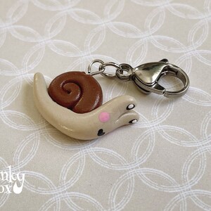 May include: A small, beige snail charm with a brown shell and pink accents. The charm is attached to a silver metal lobster clasp.