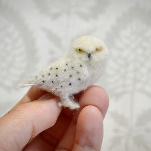 Puede incluir: Una pequeña figura de búho blanco hecha a mano, con manchas negras y ojos amarillos. El búho está hecho de un material suave y texturizado y está posado en los dedos de una persona. El fondo es un patrón floral gris claro y blanco.