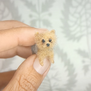 May include: A miniature Yorkshire Terrier dog figurine with tan and gray fur, black eyes, and a black nose. The small dog is held between two fingers, showcasing its tiny size. The figurine is a handcrafted item.