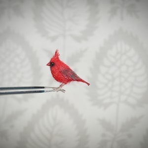 Puede incluir: Una figura de un cardenal rojo brillante posada sobre las puntas de unas pinzas plateadas. El ave tiene una textura detallada y una cresta prominente. El fondo es un papel pintado con un patrón gris claro y blanco.