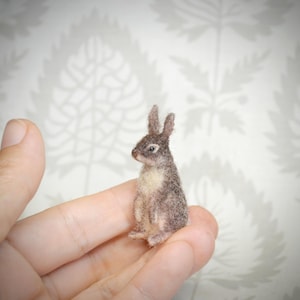 May include: A miniature needle-felted rabbit figurine featuring a textured coat of mottled brown and grey fibers with a cream-colored chest and belly. The small animal has upright ears, a dark nose, and a seated posture with visible front paws.