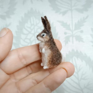 Puede incluir: Una figura de conejo en miniatura de fieltro, principalmente marrón y blanco, con orejas erguidas y un rostro detallado. La pequeña escultura se sostiene en una mano, mostrando su delicada artesanía. Un encantador ejemplo de arte animal hecho a mano.