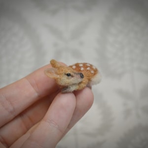 May include: A miniature felted deer figurine with a brown and white spotted body, small ears, and a black nose. The deer is lying down, and the figurine is held in a person's hand.