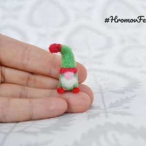 May include: A miniature felt gnome figurine. The gnome has a green body, a red hat and collar, a white beard, and a pink nose. The figurine is small, measuring approximately 2 inches in height. The text "#HromovFelts" is in the upper right corner.