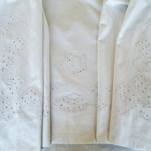 May include: Close-up of white fabric with intricate cutwork and embroidered initials. The fabric features geometric patterns and scalloped edges. The initials "JM" are embroidered in a delicate script, adding a personalized touch to the textile.