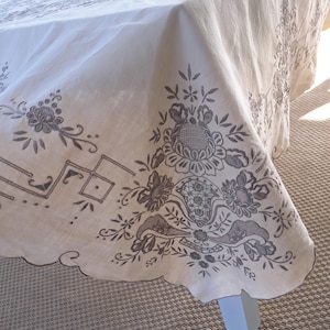 May include: A white tablecloth with a scalloped edge and intricate embroidered floral designs in brown thread.
