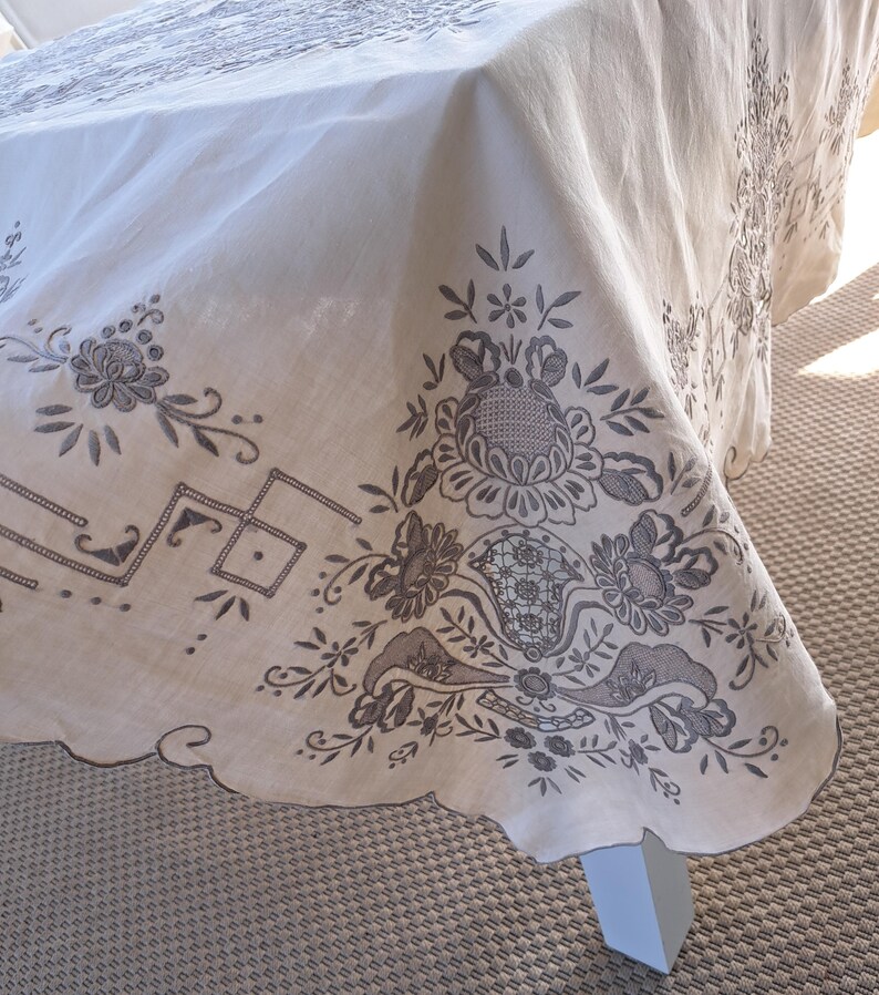 May include: A white tablecloth with a scalloped edge and intricate embroidered floral designs in brown thread.