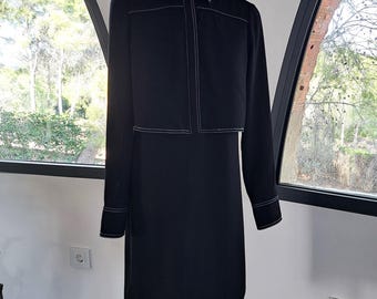 May include: A black long-sleeve shirt dress featuring a structured collar, a button-down front placket, and prominent white contrast stitching along the seams, cuffs, and hemline, displayed on a mannequin stand.