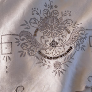 May include: A close-up of a white linen tablecloth with intricate embroidered floral designs. The embroidery is in a light gray color and features a variety of floral motifs, including leaves, flowers, and swirls.
