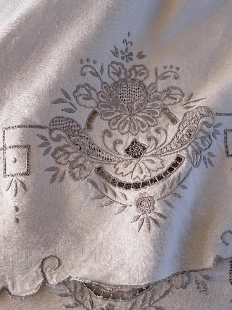 May include: A close-up of a white linen tablecloth with intricate embroidered floral designs. The embroidery is in a light gray color and features a variety of floral motifs, including leaves, flowers, and swirls.